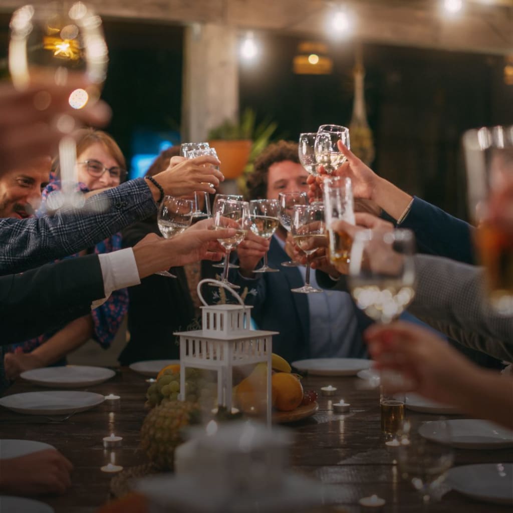 Big Dinner Party with a Small Crowd of Multiethnic Diverse Friends Celebrating at a Restaurant. Beautiful Happy Hosts Propose a Toast and Raise Wine Glasses while Sitting at a Table in the Evening.
